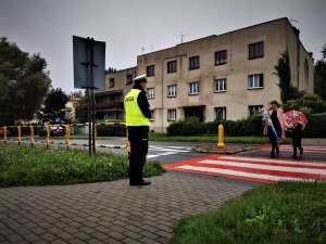 policjant ruchu drogowego w odblaskowej kamizelce stoi przy oznakowanym przejściu dla pieszych przez które przechodzą dzieci z rodziacami