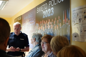 grupa seniorów na korytarzu komendy, na ścianach wiszą ogromne zdjęcia ze święta Policji, wśród nich Zastępca Naczelnika drogówki