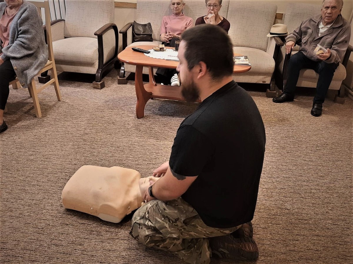 mężczyzna klęczy na dywanie i pokazuje zasady udzielania pierwszej pomocy