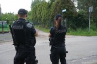 na zdjęciu widoczny patrol policji- po prawej stronie policjantka, po lewej- policjant. Oboje stoją tyłem do obiektywu i patrzą przed siebie