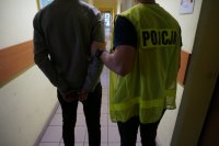 zdjęcie przedstawia policjanta w żółtej kamizelce odblaskowej z napisem &quot;policja&quot; prowadzącego mężczyznę w kajdankach