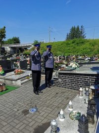 Na fotografii widać cmentarz. Przed jednym z pomników stoją dwaj policjanci i oddają honor.