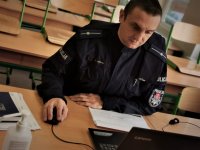 policjant siedzi w ławce szkolnej klasy, patrzy w ekran otwartego laptopa