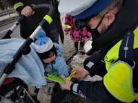 policjant drogówki przyczepia odblaskową opaskę na rączkę małego dziecka, za którym stoją rodzice