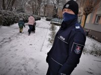 umundurowany policjant stoi przed budynkiem przedszkola w Mikołowie. W tle widać idącą z dzieckiem kobietę. Jest pełno śniegu