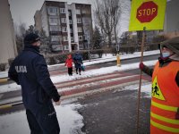 umundurowany policjant stoi przy przejściu dla pieszych. Widoczna na zdjęciu jest kobieta ze znakiem stop, która wstrzymuje ruch, by przez pasy mogli przejść piesi. Scena ma miejsce w centrum miasta. Jest poranek, chodniki są zaśnieżone.