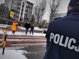 umundurowany policjant stoi przy przejściu dla pieszych. Widoczna na zdjęciu jest kobieta ze znakiem stop, która wstrzymuje ruch, by przez pasy mogli przejść piesi. Scena ma miejsce w centrum miasta. Jest poranek, chodniki są zaśnieżone.