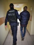 umundurowany policjant prowadzi zatrzymanego mężczyznę. Człowiek ten ma założone na rękach kajdanki