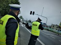 dwóch policjantów w odblaskowych kamizelkach stoi przy drodze i kontroluje prędkość
