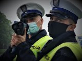 dwóch policjantów w odblaskowych kamizelkach stoi przy drodze i kontroluje prędkość, jeden z nich trzyma ręczny miernik prędkości