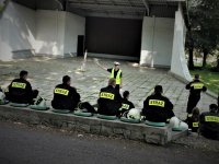 strażacy - ochotnicy siedzą na ławkach, przed nimi stoi policjant