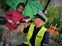 policjant ruchu drogowego, trzyma w ręku mikrofon, zadaje dziewczyne pytanie. Dziewczynka trzyma parasol