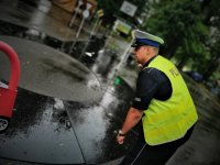 policjant drogówki wskazuje dzieciom kierunek ruchu samochodzików, pada deszcz.
