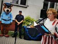 kobieta w okularach czyta do mikrofonu, w tle policjant i policyjna maskotka