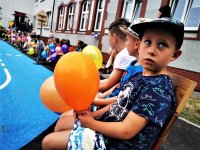 siedzące na ławce dzieci które trzymają balony, w tle budynek szkoły