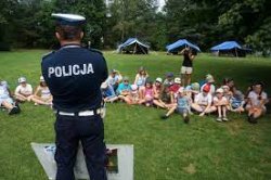 Umundurowany policjant stojący na trawie podczas spotkania z dziećmi. Na drugim planie grupa dzieci siedząca na trawie.