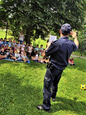 policjant prowadzi zajęcia. Grupa dzieci siedzi na trawie i słucha