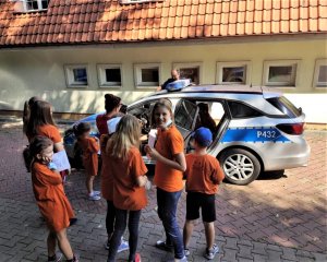 grupa dzieci w pomarańczowych koszulkach pozuje do zdjęcia. W tle policyjny radiowóz