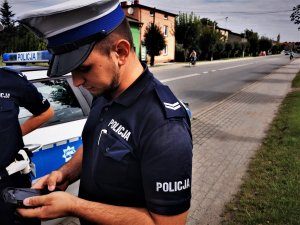 policjant drogówki stoi obok radiowozu i dokonuje sprawdzenia w urządzeniu