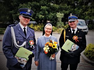 Zastępca Komendanta Policji pozuje do zdjęcia z zakonnicą oraz komendantem straży pożarnej. Kobieta trzyma bukiet kwiatów