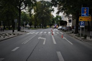 widok na jezdnię w centrum Mikołowa, widać także policyjny radiowóz oraz biały samochód osobowy. Przechodnie idą chodnikiem