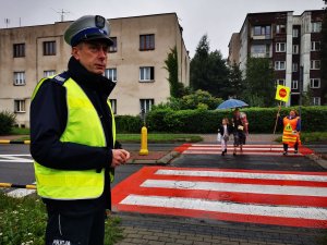 policjant ruchu drogowego w odblaskowej kamizelce stoi przy oznakowanym przejściu dla pieszych przez które przechodzą dzieci z rodziacami, widać tak zwaną panią STOPKĘ