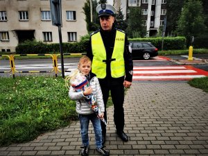 uczeń pierwszej klasy pozuje do zdjęcia z policjantem drogówki, w tle oznakowane przejście dla pieszych