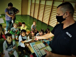 policjant w maseczce na twarzy rozdaje uczniom plany lekcji z wizerunkiem policyjnej maskotki