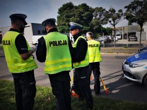 czworo policjantów drogówki w odblaskowych kamizelkach, obok fragment policyjnego radiowozu