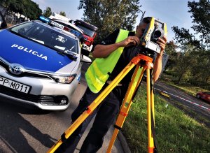 policjant drogówki w odblaskowej kamizelce pracuje na drodze, obsługuje tachimetr, za nim radiowóz