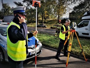 policjant drogówki w odblaskowej kamizelce pracuje na drodze, obsługuje tachimetr, obok niego drugi policjant, w tle radiowóz