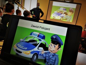 zbliżenie na monitor laptopa, na którym widać policjanta przy radiowozie. W tle siedzące dzieci oraz dzielnicowy prowadzący spotkanie