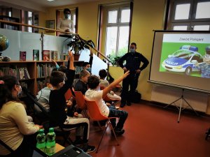 scena ma miejsce w budynku biblioteki, widać siedzących na krzesłach uczniów, przed nimi stoi policjant i omawia przygotowaną przez sienie prezentację