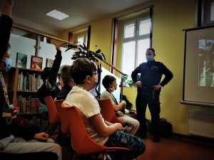 scena ma miejsce w budynku biblioteki w centrum Łazisk Górnych, widać siedzących na krzesłach uczniów, przed nimi stoi policjant i omawia przygotowaną przez sienie prezentację, która wyświetla się na rzutniku