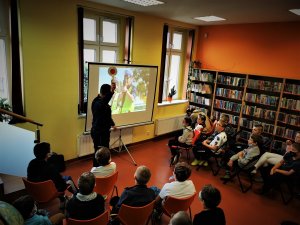 scena ma miejsce w budynku biblioteki, widać siedzących na krzesłach uczniów, przed nimi stoi policjant i omawia przygotowaną przez sienie prezentację