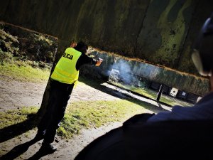 oddający strzał do tarczy policjant, za nim stoi instruktor, widać unoszący się w powietrzu dym