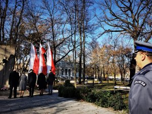 zbliżenie na fragment policyjnego munduru, w drugim planie widać pomnik i powiewające flagi