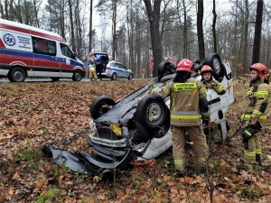 samochód leży w rowie, wokół niego stoją strażacy, w tle pojazd karetki oraz policji