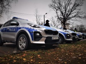 ustawione w rzędzie na parkingu trzy samochody oznakowane marki KIA, przy każdym z nich stoi policjant