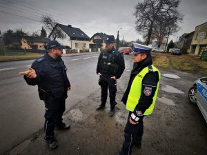 trzech policjantów stoi przy głównej ulicy miejscowosci, rozmawiają ze sobą, w tle widać budynki mieszkalne