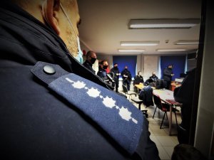 zbliżenie na policyjny pagon, w tle sala odpraw, na której siedzą umundurowani policjanci. Wszyscy mają założone maseczki ochronne