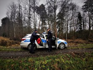 policjanci wraz z zatrzymanym wsiadają do oznakowanego samochodu, zaparkowanego w lesie, zatrzymany ma czerwoną kurtkę