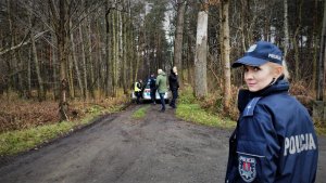 patrząca w obiektyw aparatu policjantka, w tle wejście do lasu, policyjny radiowóz oraz stojący obok niego policjanci