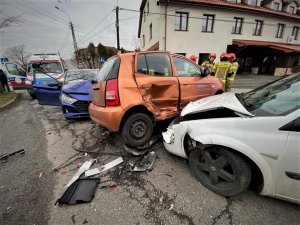 trzy rozbite podczas kolizji drogowej samochody osobowe: biały, pomarańczowy i niebieski, z tyłu budynki