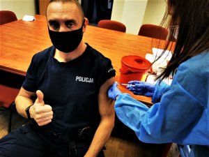 lekarka w niebieskim kitlu szczepi policjanta. Mężczyzna unosi kciuk do góry