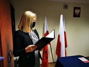 kobieta w blond włosach trzyma otwartą teczkę, w środku ma tekst, za nią 3 flagi Polski