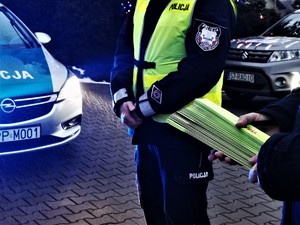 policjant w odblaskowej kamizelce, fragment radiowozu, samochodu radia oraz trzymany przez strażaka plik odblaskowych opasek