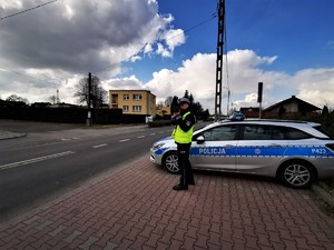 policjant drogówki w odblaskowej kamizelce stoi przy drodze i dokonuje pomiaru prędkości pojazdów. W tle policyjny radiowóz