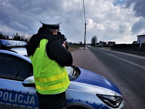 policjant drogówki w odblaskowej kamizelce stoi przy drodze i dokonuje pomiaru prędkości pojazdów. W tle policyjny radiowóz