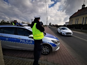 policjant drogówki stoi w kamizelce odblaskowej i dokonuje pomiaru prędkosci jadących samochodów